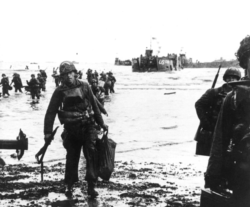 Carrying a full equipment, American assault troops move onto Utah Beach on the norther coast of France. Landing craft, in the background, jams the harbor.